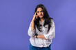 © Daniel - A young woman of Indian descent, wearing a white striped shirt and black smartwatch, poses thoughtfully with her finger on her temple, thought against a solid purple background