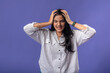 © Daniel - A young woman of Indian descent, wearing a white striped shirt and black smartwatch, stands against a solid purple background, expressing frustration by holding her head with both hands