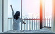 © Chinnapong - Hotel relaxation on lazy day morning with Asian woman waking up from good sleep, resting in comfort bedroom looking toward sea city view with happy work-life balance living quality luxury lifestyle