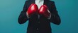 © Ai Studio - A businessperson in a dark suit poised for action, wearing red boxing gloves symbolizing readiness and competitive spirit against a blue background.