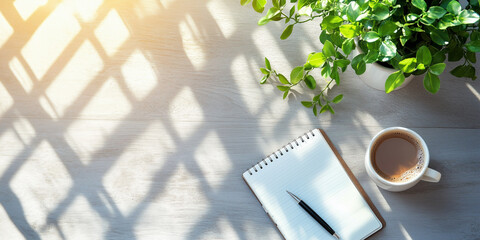 Blank notebook page with pen coffee cup and plant on wooden desk receiving sunlight from window