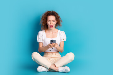 Poster - Full length photo of lovely young lady sit floor gadget frightened dressed stylish white garment isolated on blue color background