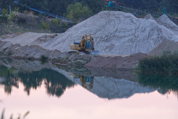 At dusk, an excavator stands above the water in a gravel pit, with a mountain of extracted gravel in the background.