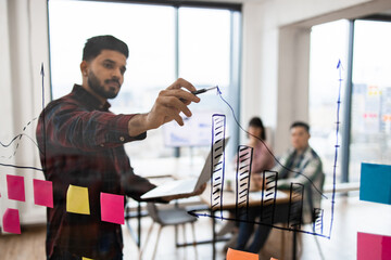 Wall Mural - Business professional presenting data analysis on transparent board during meeting. Team collaboration with bar charts and sticky notes in modern office setting.