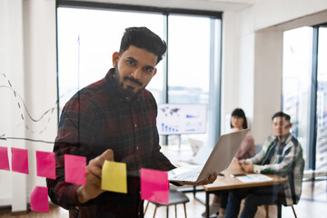 Wall Mural - Business team brainstorming session with sticky notes on transparent board. Mid-adult man leading discussion with laptop while colleagues observe from table. Modern office setting.