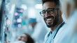 © Maximages  - A smiling male doctor wearing glasses and scrubs, standing confidently in a clinical setting with medical equipment in the background, exuding professionalism.