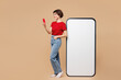 © ViDi Studio - Full body side view young woman wear red t-shirt casual clothes big huge blank screen mobile cell phone with area using smartphone isolated on plain pastel light beige background. Lifestyle concept.