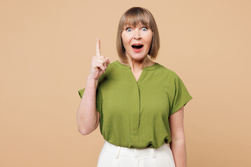 Wall Mural - Elderly blonde woman 50s years old she wear green shirt casual clothes holding index finger up with great new idea isolated on plain pastel light beige background studio portrait. Lifestyle concept.