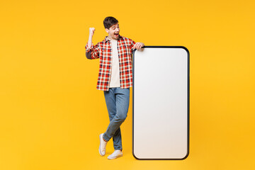 Sticker - Full body young man he wears red checkered shirt casual clothes big huge blank screen mobile cell phone smartphone with area do winner gesture isolated on plain yellow background. Lifestyle concept.