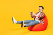 © ViDi Studio - Full body gambling young man he wears red checkered shirt casual clothes sit in bag chair hold in hand play pc game with joystick console isolated on plain yellow orange background. Lifestyle concept.