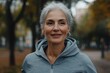 © Milos - An elderly woman with gray hair enjoys a moment in a park, dressed in a gray hoodie and exhibiting a cheerful smile, highlighting fitness and well-being in her older age.
