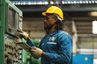© KANGWANS - A man in a blue jacket is wearing a yellow hard hat and pointing at a computer screen. He is wearing gloves and he is working in a factory