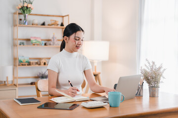 Wall Mural - A young woman is focused on her work at stylish desk, writing notes and using laptop in bright, modern home office. atmosphere is calm and productive, perfect for creativity.