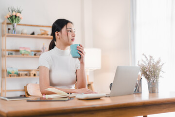 Wall Mural - A woman sits at wooden desk, sipping from blue mug while working on laptop. cozy workspace features plants and decorative items, creating warm atmosphere.