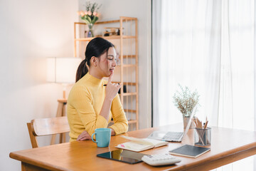 Wall Mural - A woman in yellow sweater sits thoughtfully at wooden desk, surrounded by cozy workspace. She gazes at her laptop, with cup of coffee and stationery nearby, creating serene atmosphere.