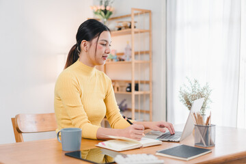 Wall Mural - A woman in yellow sweater is working on her laptop at cozy desk, surrounded by plants and stationery, creating productive and inviting atmosphere.