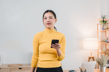 Wall Mural - A young woman in yellow turtleneck sweater stands confidently holding smartphone in modern workspace, exuding sense of professionalism and focus.