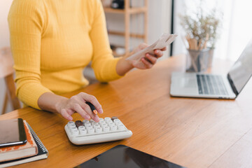 A person in yellow sweater is using calculator while holding receipts, surrounded by laptop and notebooks, creating focused and productive atmosphere.