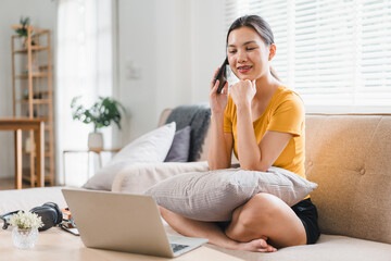 Wall Mural - A young woman is sitting comfortably on sofa, engaged in phone conversation while using laptop. She appears relaxed and happy, surrounded by cozy living space filled with natural light.