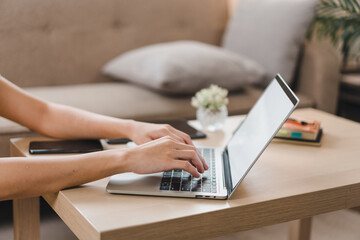 Wall Mural - A person is working on laptop at wooden table in cozy living room, surrounded by soft furnishings and plants, creating productive and inviting atmosphere.