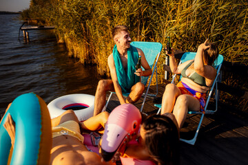 Wall Mural - Group of friends in swimsuit having fun sitting on a pier drinking beer enjoying a summer day at the lake.