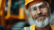 © Maximages  - An older bearded construction worker wearing a yellow helmet and protective glasses smiles warmly, standing on-site, symbolizing experience, dedication, and job satisfaction.