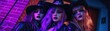 © nattapon - Three women in gothic attire and hats, illuminated by purple and blue neon lights, and standing against a brick wall