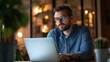 © fotofabrika - Man working late at night on a laptop in a cozy, well-lit home office filled with plants and ambient lighting