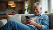 © Ali Hamza Tullah - Relaxed happy older mature middle aged man holding mobile phone using cellphone sitting at home on sofa in modern living room, chatting online, texting messages, reading news, shopping in apps