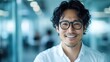 © Maximages  - A smiling man with curly hair and glasses, wearing a white shirt, stands in a bright, modern office setting, captured in a friendly and professional manner.