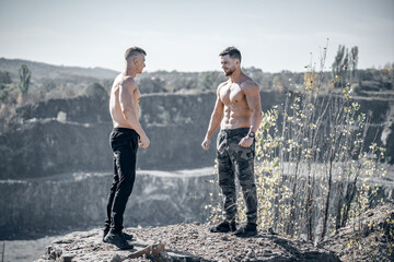 Two shirtless men engage in a flexing competition outdoors. Two muscular men stand on a rocky ledge, flexing their biceps in a friendly competition amidst a natural landscape.