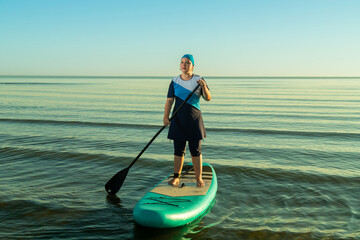 Woman in Muslim burkini paddling on SUP board in sea against sunrise sky