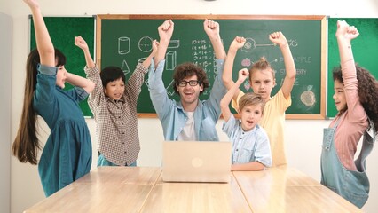 Wall Mural - Teacher and multicultural student looking at laptop while celebrate success project and put hands in the air. Group of children smiling to camera while standing at blackboard in classroom. Pedagogy.