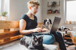 © VisualProduction - A woman sitting on a wooden bench, working on a laptop. A black cat is lying on her lap, and a dog is sitting beside her, looking at the camera. The room has plants and cozy decor.