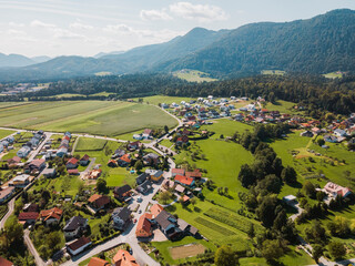 Aerial view of a picturesque village surrounded by lush green fields and mountains. The landscape features a mix of residential homes, winding roads, and agricultural land, showcasing the beauty of ru