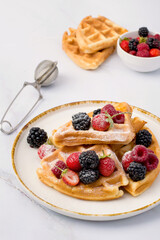 Belgian waffles decorated with strawberries, raspberries, blackberries and sprinkled with powdered sugar on a light background.