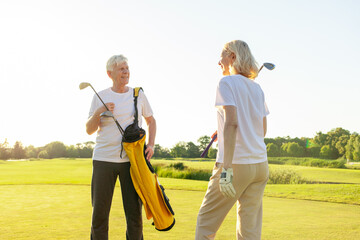 old senior couple in white clothes playing golf on golf course and chatting at sunset, elderly man and woman actively relaxing outdoors and laughing, pensioners on vacation