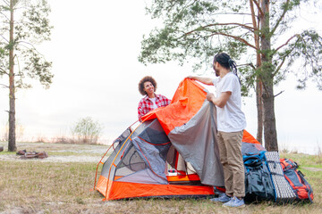 young african american couple hikers pitching tent in forest near lake, man and woman with camping equipment and backpacks traveling and preparing camping site