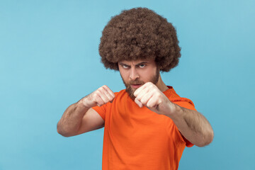 Portrait of man with Afro hairstyle in orange T-shirt aggressive fighter holding clenched fists up ready to boxing, martial art trainer, self defense. Indoor studio shot isolated on blue background.