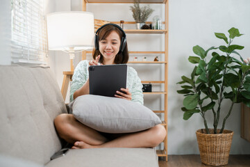 a young woman sitting on a sofa, using a tablet and wearing headphones in a cozy, modern living room