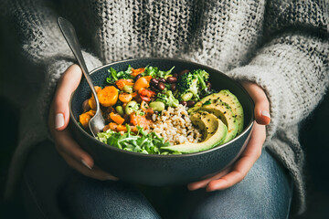 Healthy vegetarian dinner. Woman in jeans and warm sweater holding bowl with fresh salad, avocado, grains, beans, roasted vegetables, close-up. Superfood, clean eating, vegan, dieting food   