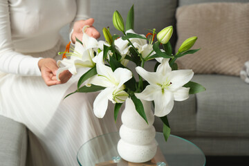 Wall Mural - Beautiful young African-American woman with vase of bouquet of white lily flowers sitting on sofa at home, closeup