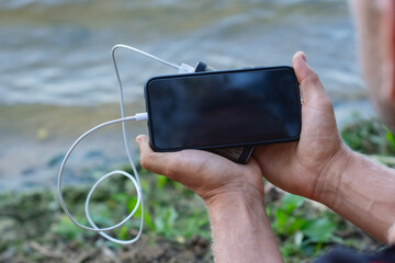 The guy is holding a portable charger with a smartphone in his hand. Man on a background of nature with a greenery and a lake