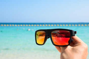 Hand holding a cool black sunglasses with orange lenses on a tropical beach and blue sea background morning day at Koh Larn island. Summer and travel concept. Vacation. Pattaya, Thailand.