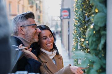 Wall Mural - Happy couple enjoys the festive season shopping in the city streets filled with christmas lights and decorations, laughing and looking at holiday displays while carrying bags