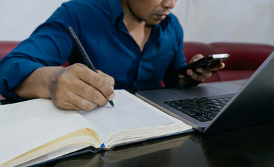 Business planning, Elearning concept. Casual business man writing on notebook and working on laptop computer at home. Man using mobile phone, studying online course via laptop and lecture on notepad.