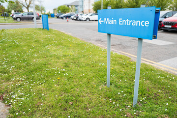 Shallow focus of a generic, blue Main Entrance sign seen at a typical NHS hospital. The sign navigates patients to the main out patients building.