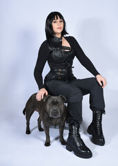 Wall Mural - Full length portrait of beautiful black female model wearing sci fi futuristic dystopian leather bodysuit costume, with animal dog pet companion. sitting pose, isolated white studio background.