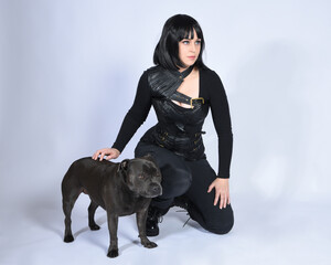 Wall Mural - Full length portrait of beautiful black female model wearing sci fi futuristic dystopian leather bodysuit costume, with animal dog pet companion. sitting pose, isolated white studio background.