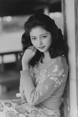 A Japanese woman seated on a modern bench, her right hand resting on her knee while her left hand adjusts the sleeve of her sweater.